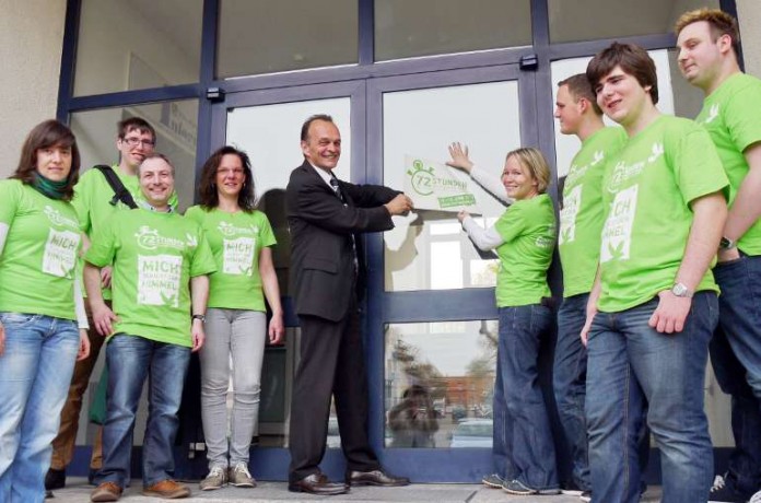 Landrat Dr. Fritz Brechtel u. Susanne Poerschke, Jugendreferentin für das Dekanat Germersheim, bringen ein 72-Stunden-Aktion-Logo auf der Eingangstür der Kreisverwaltung Germersheim an. Mit dabei war auch das Koordinationsteam